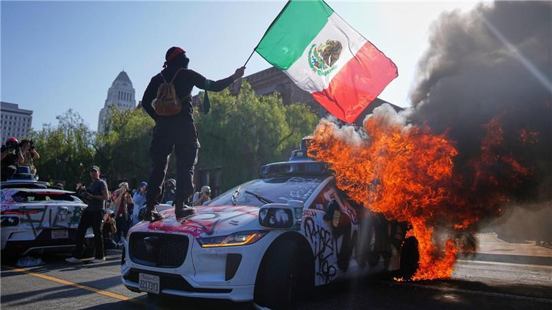 Ein Demonstrant steht auf einem brennenden Waymo-Taxi in der Nähe des Metropolitan Detention Center in der Innenstadt von Los Angeles.