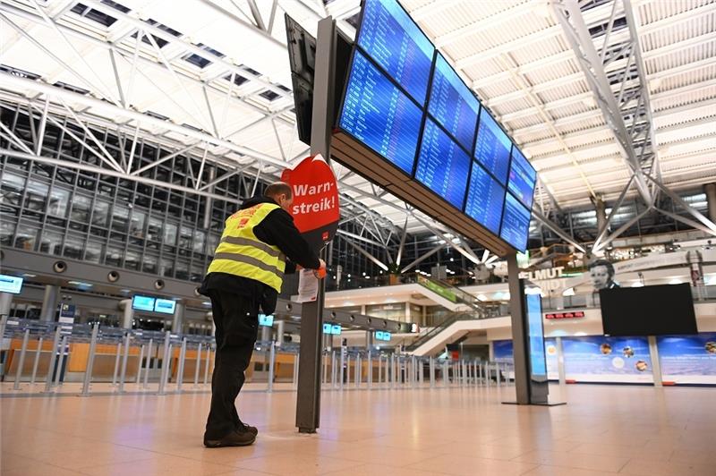 Warnstreik: Gähnende Leere am Hamburger Flughafen Ein Gewerkschaftsvertreter klebt ein Plakat mit der Aufschrift "Warnstreik!" und einen Flyer an die Anzeigetafel im leeren Terminal 2. Am Hamburger Flughafen hat am Sonntagabend ein Warnstreik von Beschäftigten begonnen. Foto: Jonas Walzberg/dpa