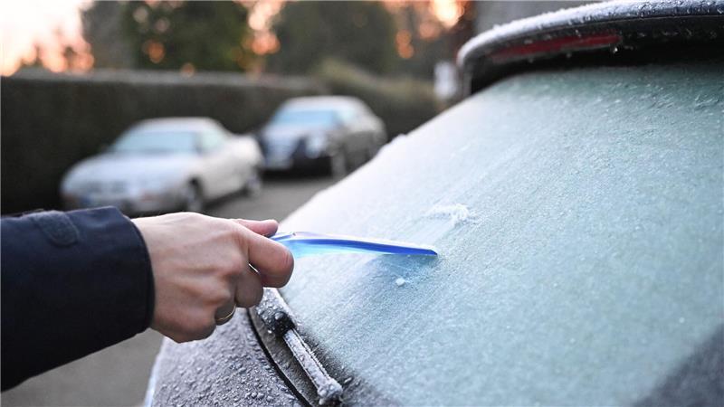 Ein Mann kratzt am Morgen eine Autoscheibe von einer Eisschicht frei.
