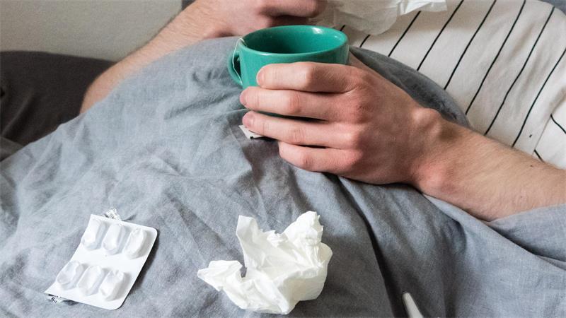 Ein Mann liegt mit Taschentüchern, Teetasse, Nasenspray und Tabletten im Bett (gestellte Szene). 