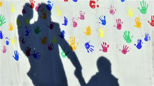 Ein Mann mit einem Kind auf dem Arm und einem an der Hand wirft einen Schatten auf eine mit bunten Handabdrücken bemalte Wand einer Kindertagesstätte. (Symbolbild)