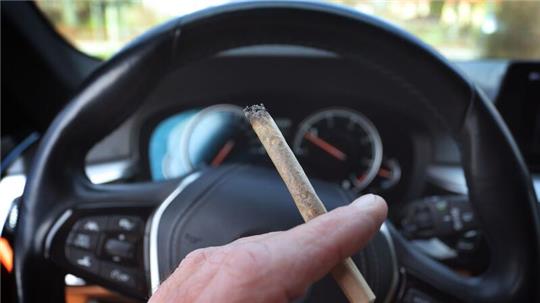 Ein Mann sitzt mit einem Joint zwischen den Fingern am Steuer eines Autos.