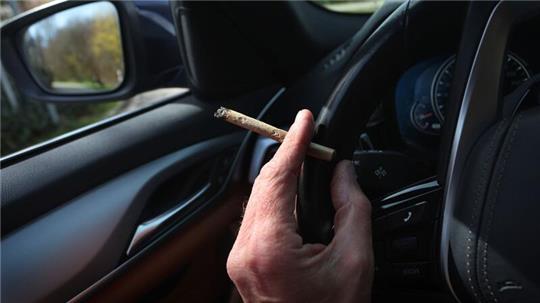 Ein Mann sitzt mit einem Joint zwischen den Fingern am Steuer eines Autos.