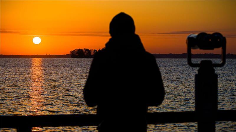Ein Mann steht im Licht der aufgehenden Sonne neben einem Fernglas an einem Aussichtspunkt am Steinhuder Meer in der Region Hannover.