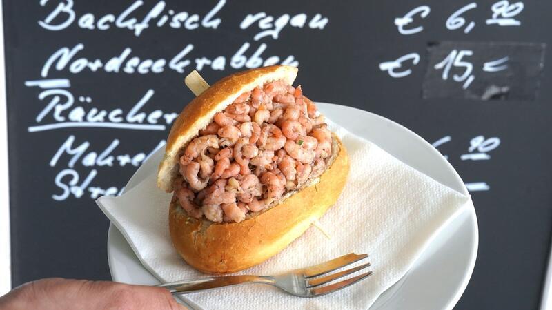 Wird das Krabbenbrötchen zum Luxusgut? Ein Mitarbeiter hält ein Nordseekrabben-Brötchen vor einer Preistafel „Nordseekrabben - 15,- Euro“.