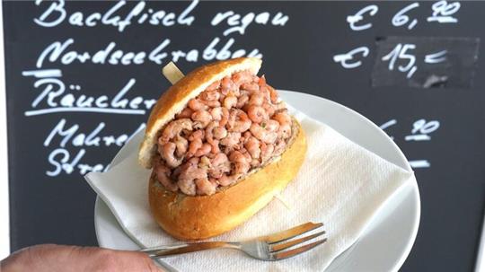 Ein Mitarbeiter hält ein Nordseekrabben-Brötchen vor einer Preistafel „Nordseekrabben - 15,- Euro“.