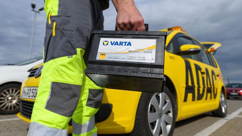 Ein Pannenhelfer der ADAC-Straßenwacht hält vor seinem ADAC Auto eine Autobatterie.