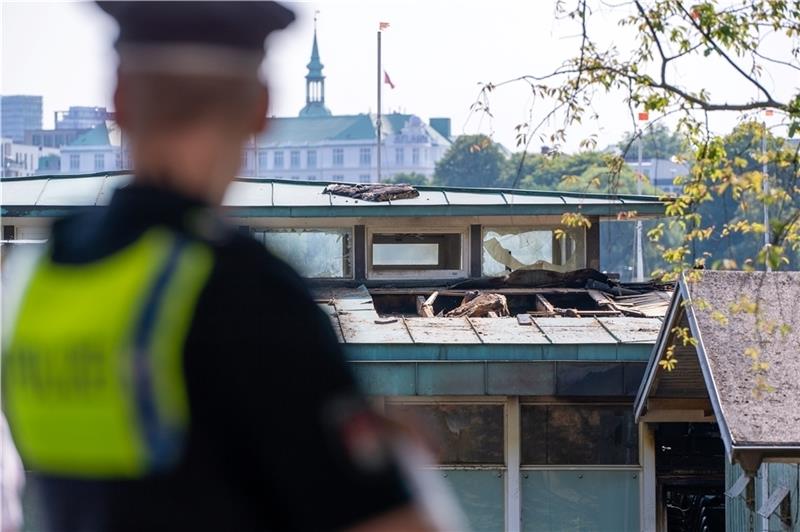 Ein Polizist steht nahe des aufgetrennten Dachs eines bekannten Gastronomiebetriebs eines Segelclubs an der Hamburger Außenalster, in dem am Freitagmorgen ein Feuer ausgebrochen ist. Foto: Jonas Walzberg/dpa