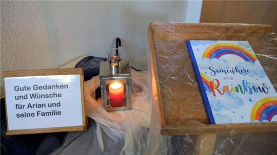 Ein Schild steht neben einer Kerze und ein Buch mit einem Regenbogen auf dem Umschlag liegt daneben in der Kirche der Gemeinde Elm.