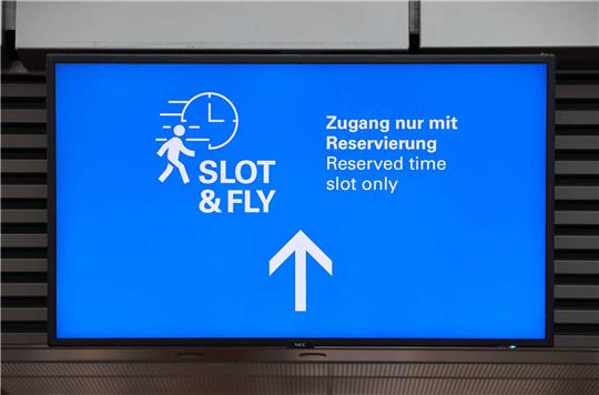 Ein Screen mit der Aufschrift "Slot &amp;Fly  - Zugang nur mit Reservierung" ist am Hamburger Flughafen zu sehen.  Jeder Passagier hat künftig die Möglichkeit, sich kostenlos einen 15-Minuten-Zeitslot für einen separaten Zugang zur Sicherheitskontrolle zu reservieren. Foto: Georg Wendt/dpa