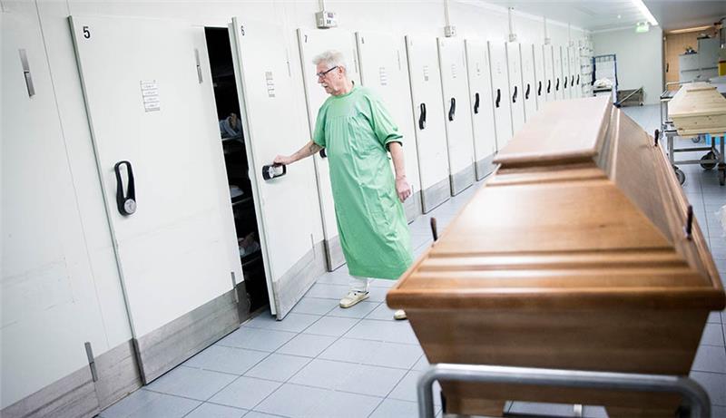 Ein Sektionsassistent öffnet im Institut für Rechtsmedizin in Hamburg die Tür zu einem Leichenkühlfach neben den Sektionsräumen im Keller des Instituts. Neben ihm steht eine Bahre mit Holzsarg (Symbolbild). Foto: Christian Charisius/dpa