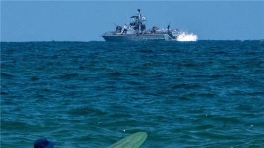 Ein Surfer wartet auf eine Welle, während ein israelisches Marineschiff im Mittelmeer vor der Küste von Hadera patrouilliert.