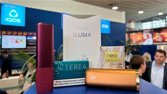 Ein Tabakerhitzer der Marke Iqos vom Hersteller Philip Morris und ein dazugehöriges Päckchen Tabaksticks der Marke Terea sowie das Konkurrenzprodukt Glo (mit Neo-Tabaksticks) des Herstellers BAT liegen bei der Fachmesse Intertabac in einer Messehalle.