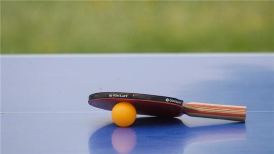 Ein Tischtennisschläger und ein gelber Tischtennisball liegen auf einer blauen Tischtennisplatte.