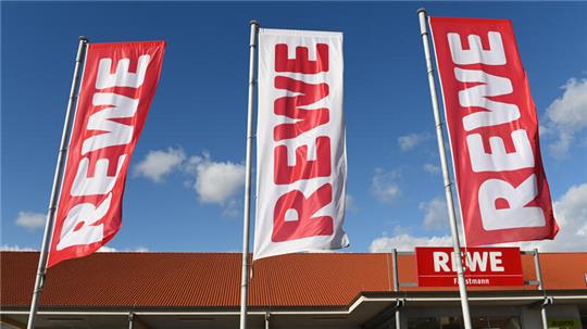 Fahnen mit dem Logo des Lebensmitteleinzelhändlers Rewe wehen vor einer Filiale.