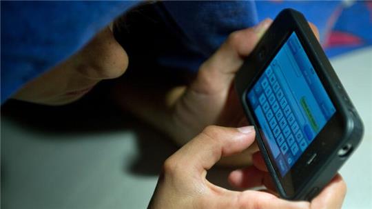 Ein elfjähriger Junge liegt unter der Bettdecke und schreibt auf einem Smartphone eine SMS. Kinder unter 13 Jahren sollten Experten zufolge keinen Zugang zu derartigen Medien haben.