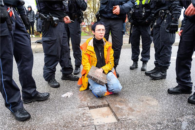 Eine Aktivistin sitzt mit einem Betonteil an ihrer Hand zwischen Polizisten auf der Fahrbahn. Klimaaktivisten der Bewegung "Letzte Generation" haben am 25. März die Elbbrücken stadteinwärts blockiert. Neben Strafanzeigen will die Hamburger Polizei auch die Kosten des Einsatzes und der Fahrbahnreparatur geltend machen. Foto: Jonas Walzberg/dpa