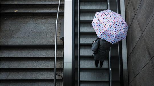Eine Frau fährt mit ihrem Regenschirm eine Rolltreppe hinauf. Das Wetter soll in der Region Stuttgart in den nächsten Tagen wechselhaft bleiben.