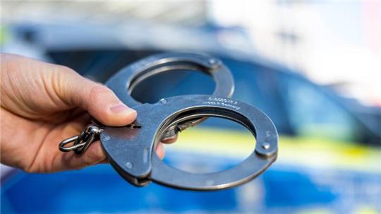 Eine Hand hält Handschellen vor einen Streifenwagen der Polizei.