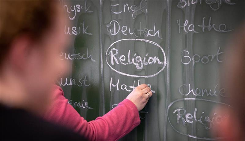 Eine Lehrerin schreibt mit Kreide einen Stundenplan mit Religionsunterricht an eine Tafel. Foto: Friso Gentsch/dpa