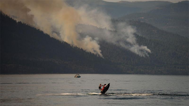 Eine Person fährt mit einem Jetski, währen im Hintergrund ein Wald brennt.