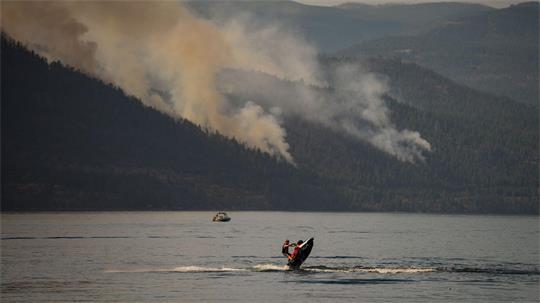 Eine Person fährt mit einem Jetski, währen im Hintergrund ein Wald brennt.