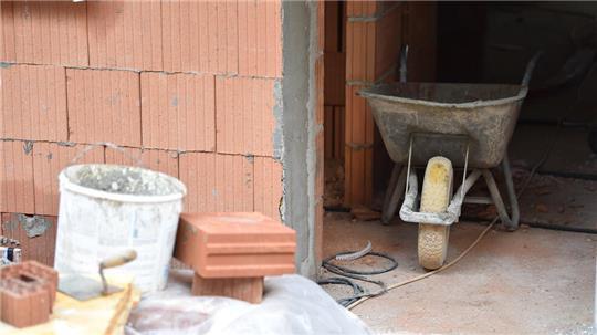 Eine Schubkarre, Steine, Maurerkelle und ein Mörteleimer stehen auf einer Baustelle.