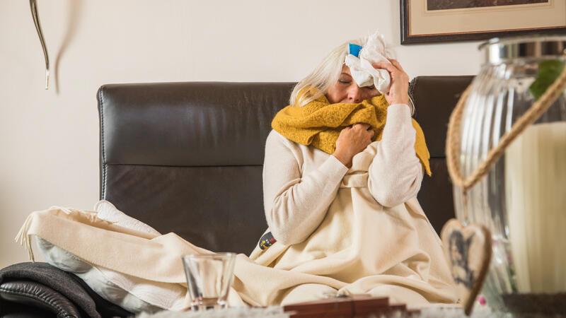 Eine aeltere Frau sitzt krank auf ihrer Couch und kuehlt ihre Stirn (gestellte Szene).