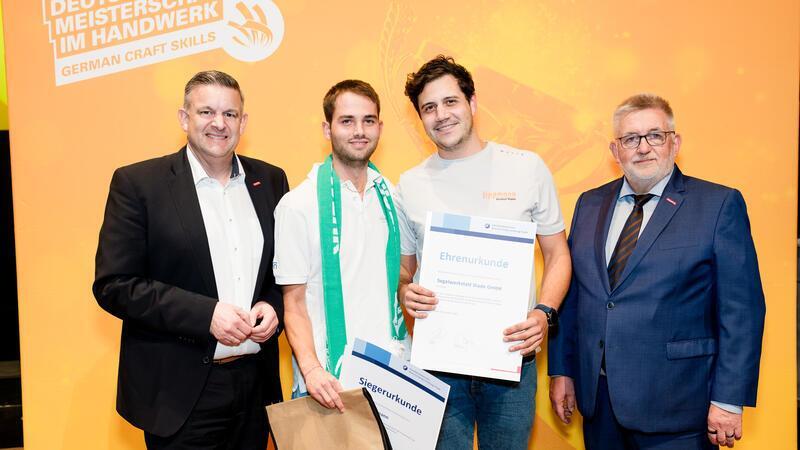 Matti Pleßmann im weißen Poloshirt steht mit einer Siegerurkunde in der rechten Hand neben seinem Ausbilder und zwei Herren im Anzug.