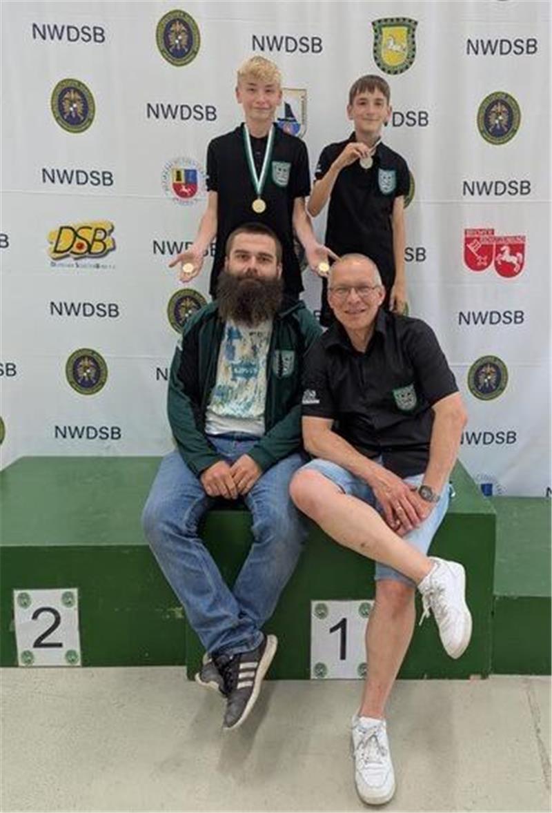 Die Trainer Thoralf Lemmel und Ralf Richter sitzen vorne auf einem Siegerpodest, dahinter stehen ihre Schützen Nelian Gehlken und Lian Eickstädt.