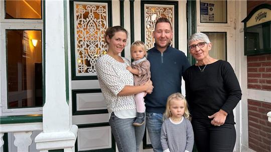 Eine junge Familie steht vor der verzierten Eingangstür eines Gasthauses und neben einer hölzernen Säule. 