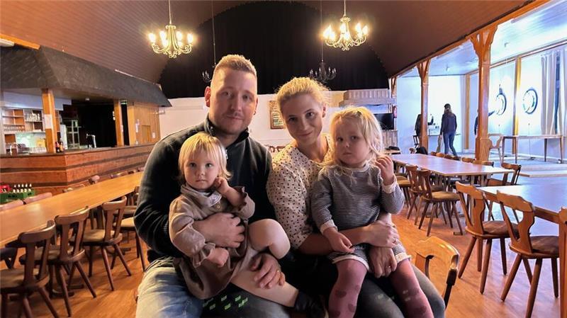 Gasthaus als Familiensache: Neustart im Großenwördener Hof Ein junges Ehepaar, Felix und Katharina Fitze, hat zwei kleine Mädchen auf dem Schoß. Im Hintergrund sind Tische und Stuhlreihen in einem großen Saal zu sehen.