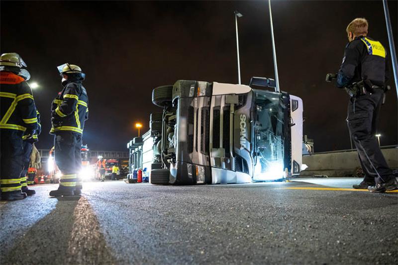 Feuerwehrleute und ein Polizist stehen neben dem umgekippten Sattelzug auf der Autobahn 7. Vor dem Hamburger Elbtunnel ist am Mittwochabend ein Lastwagen umgekippt. Der Fahrer wurde dabei leicht verletzt, wie ein Polizeisprecher sagte. Foto: Jonas Walzber
