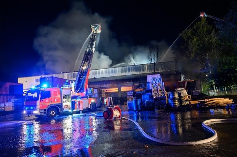 Flammenschein ist in einer Lagerhalle zu sehen, während zwei Drehleiterfahrzeuge von außen löschen. Foto: Jonas Walzberg/dpa