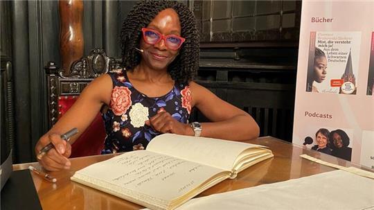 Florence Brokowski-Shekete sitzt am Holztisch im historischen Ratssaal mit dem aufgeschlagenen Buch samt vollendetem Eintrag vor sich liegend