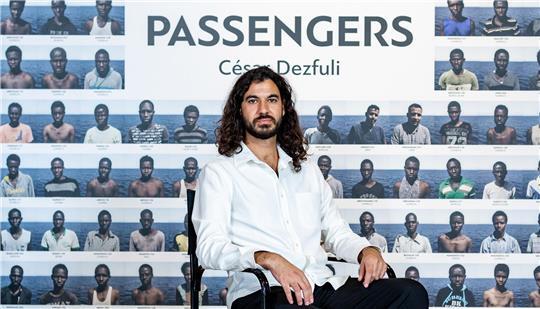Fotograf César Dezfuli vor einem Mosaik aus den 118 Ausstellungsporträts, die er 2016 an Bord des zivilen Seenotrettungsschiffes Iuventa gemacht hat.