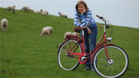 Heike Götz ist unterwegs als Fernsehmoderatorin der NDR-Landpartie und steht, umgeben von Schafen, mit ihrem roten Fahrrad am Deich