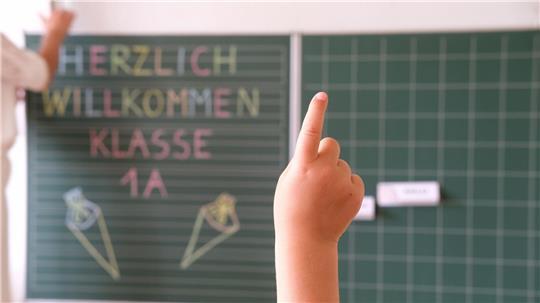 „Herzlich willkommen Klasse1 A“ steht an der Tafel eines Klassenzimmers, während sich ein Kind meldet. Am Samstag wird in Sachsen Einschulung gefeiert.