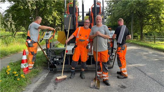 Zwei Bauarbeiter bedienen einen Aspahltfertiger, zwei stehen davor und schauen in die Kamera.