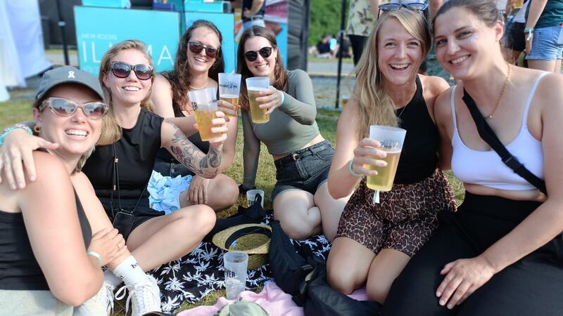 Eine Gruppe junger Frauen sitzt lachend auf der Festivalwiese und trinkt Bier.