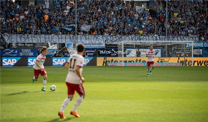 Hoffenheimer Fans halten einen Banner mit der Aufschrift "Die Liga ist euch endlich los. Der Dino kriegt den Gnadenstoß". Im Vordergrund laufen Spieler des HSV. Foto: Sebastian Gollnow/dpa