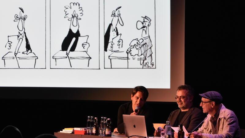 So kommt die bissige Titanic-Satire auf der Holk-Bühne an Auf einer Leinwand werden die entsprechenden Cartoons gezeigt, schräg davor sitzen Elias Hauck, Dominik Bauer und Thomas Gsella hinter einem Tisch