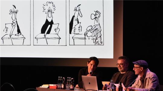 Auf einer Leinwand werden die entsprechenden Cartoons gezeigt, schräg davor sitzen Elias Hauck, Dominik Bauer und Thomas Gsella hinter einem Tisch 