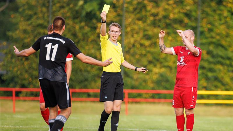 Ein Schiedsrichter zeigt die Gelbe Karte, ein Spieler verhöhnt seinen Gegner.
