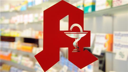 In einer Apotheke ist ein Apothekenlogo an einer Glastür angebracht. Dahinter stehen in Regalen verschiedene Medikamente. (Symbolbild)
