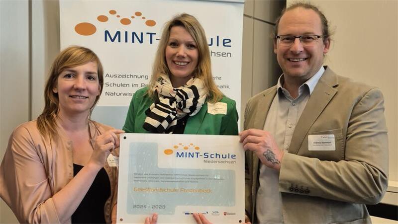 Jessica Bönsch (Nordmetall-Stiftung, links) überreicht das neue Schild an die Vertreter der Geestlandschule Daniela Bielke (MINT-Koordinatorin) und Andreas Dammert (Koordinator Berufsorientierung).