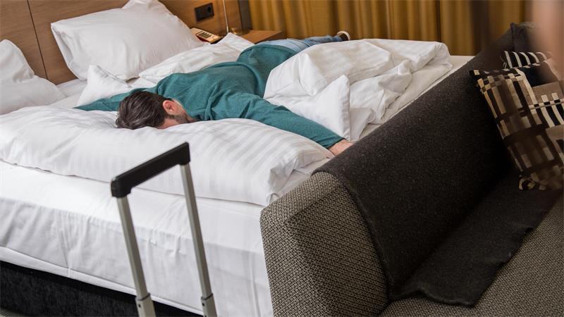 Ein Mann liegt erschöpft auf einem Bett in einem Hotelzimmer. 