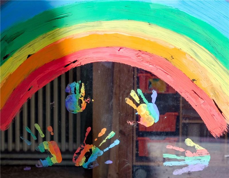 Kinderhände und ein Regenbogen am Fenster einer Kita.