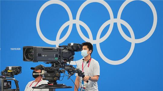 Ein TV-Kameramann steht mit der TV-Kamera am Beckenrand unter den olympischen Ringen
