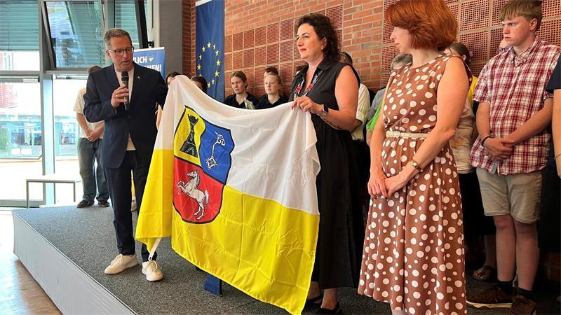 Landrat Kai Seefried, Übersetzerin Viktoria Kirn und Konsulin Olha Novytska enthüllen eine Flagge des Landkreises Stade, die – mit den Unterschriften der Helferinnen und Helfer – nach Mykolajiw geht.